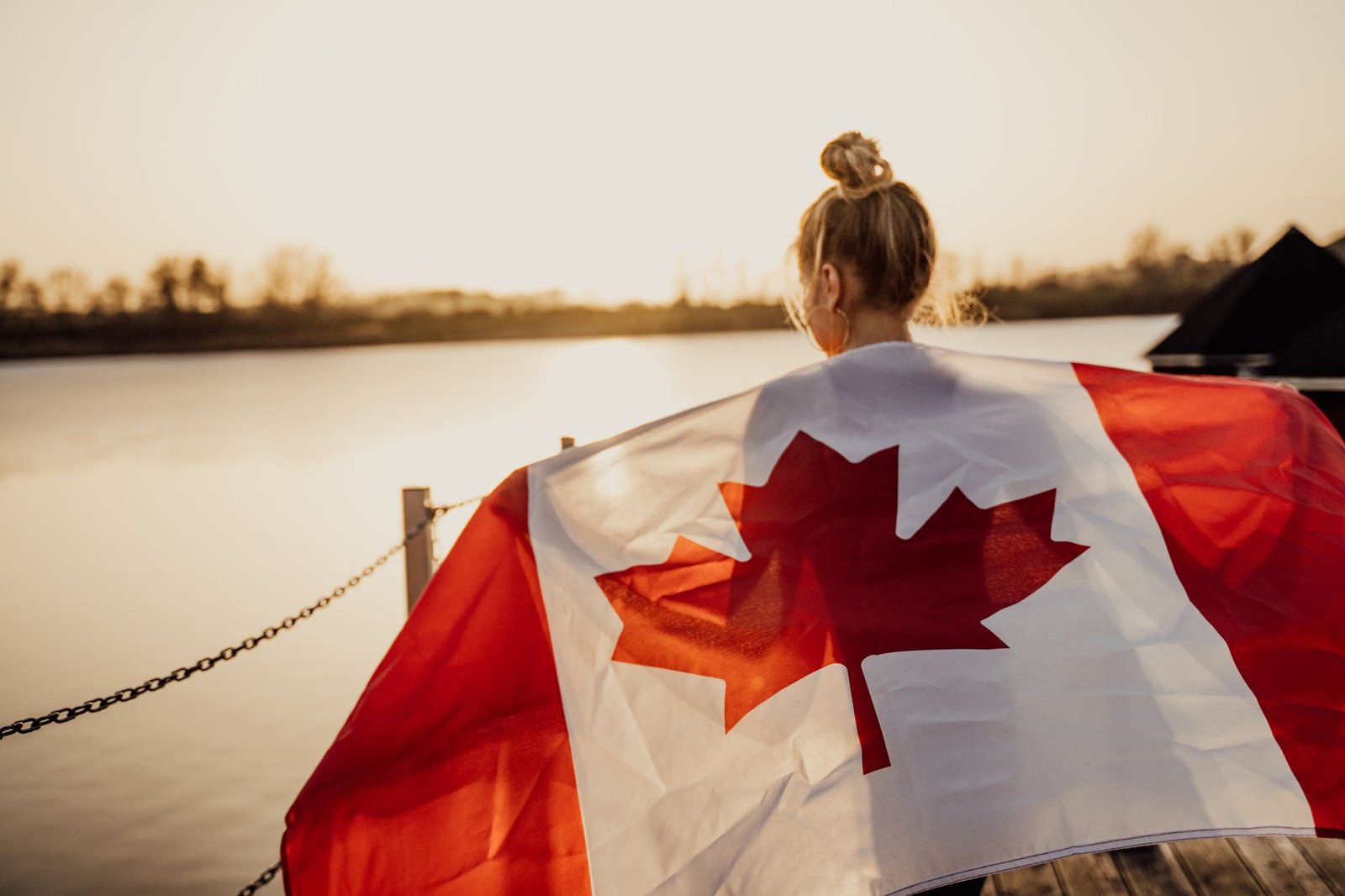 A woman holding a Canadian flag at a lakeside pier during sunset